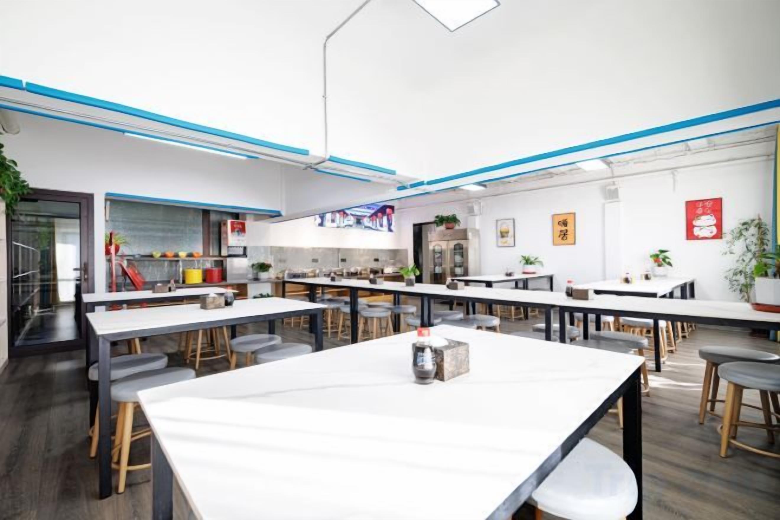 A modern dining area with long tables and chairs, a kitchen in the background, and a red sign on the wall.