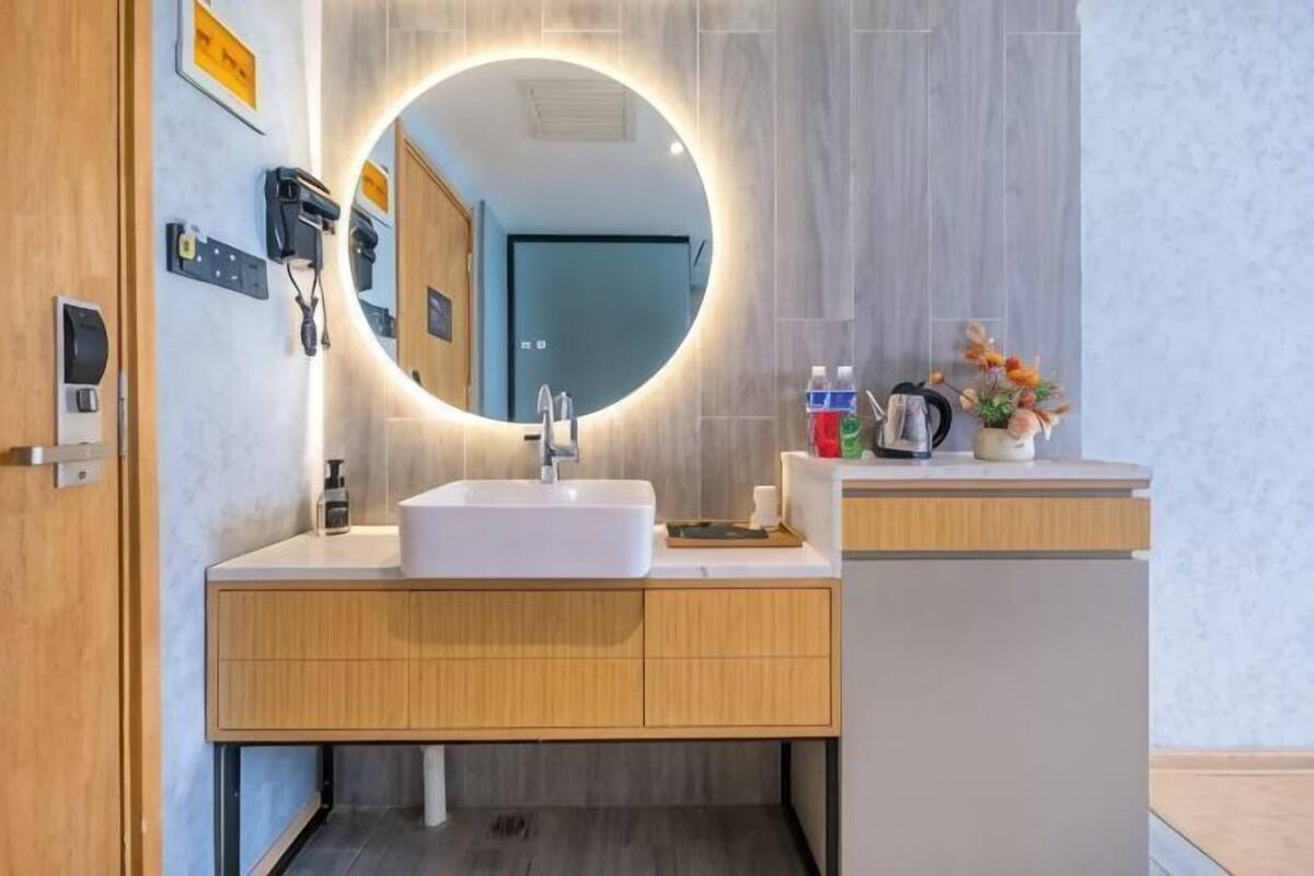 Un baño moderno con un espejo redondo, un lavamanos blanco, armarios de madera y una planta pequeña en la encimera.