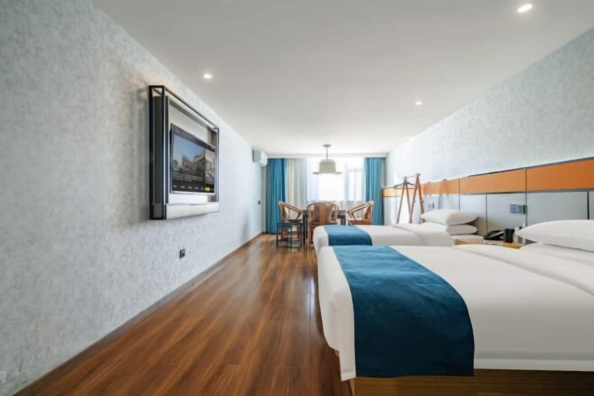 Une chambre d’hôtel avec deux lits, un plancher en bois, une grande fenêtre avec des rideaux, un luminaire suspendu et un tableau encadré au mur.