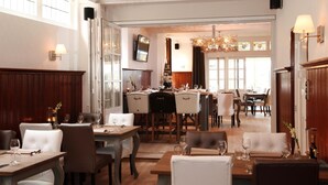 A hotel dining area with multiple tables and chairs, a chandelier, and a television mounted on the wall. - Hotel Restaurant Riche (Boxmeer)