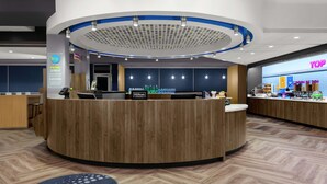 Zona de recepción de hotel con un mostrador de madera, una pantalla digital y una barra con botellas de agua.