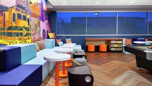 Un área de estar moderna con asientos de colores, un mural y vistas de la ciudad al anochecer.
