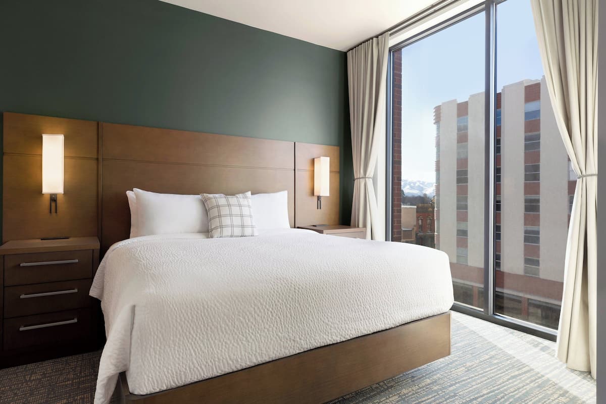 A hotel room with a large bed, two bedside tables, a nightstand, a window with curtains, and a view of a building.
