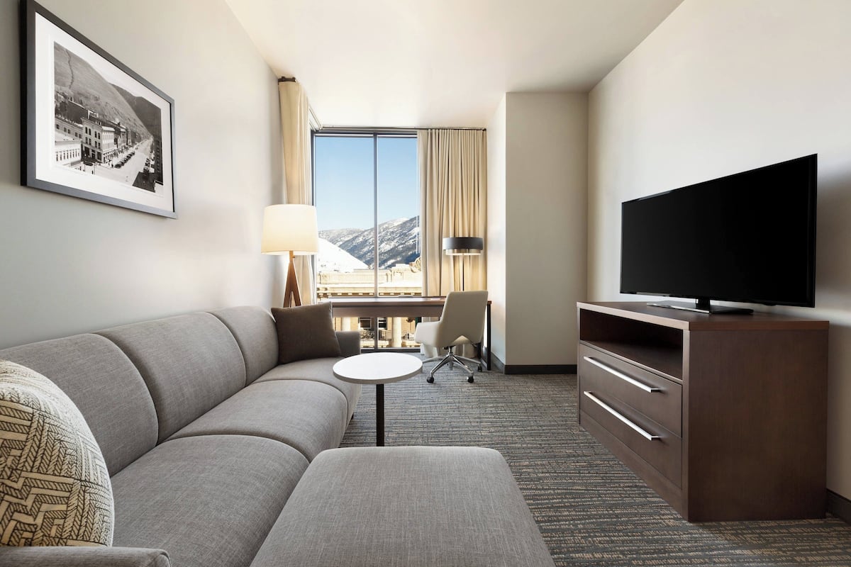 A hotel room with a sofa, a small table, a TV on a wooden stand, a desk with a chair, and a large window with a view of mountains.