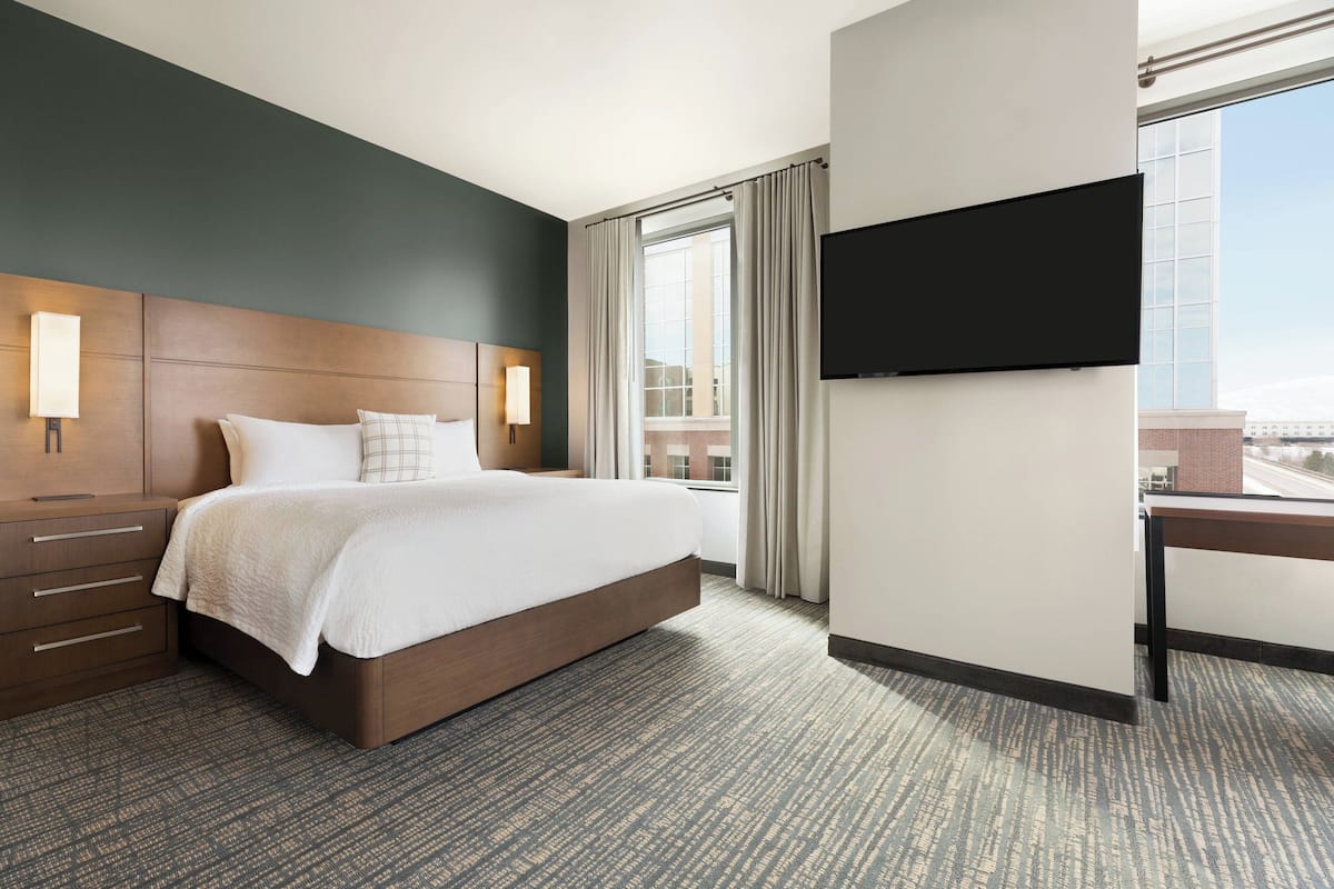A hotel room with a large bed, a flat-screen TV mounted on the wall, a window with curtains, and a view of the cityscape.