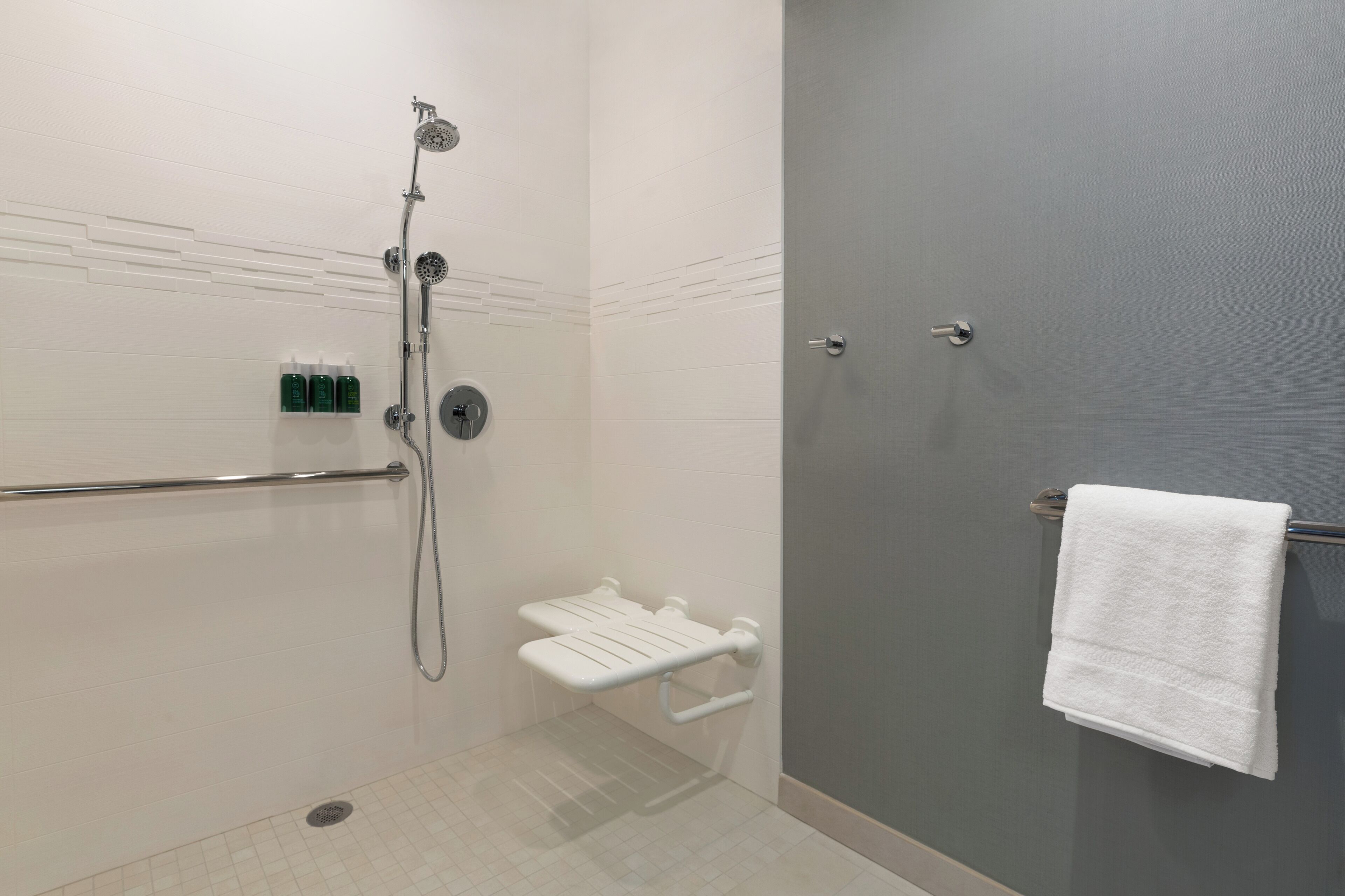 A shower area with a handheld showerhead, a handheld shower seat, a towel rack with a white towel, and a set of green bottles on the wall.