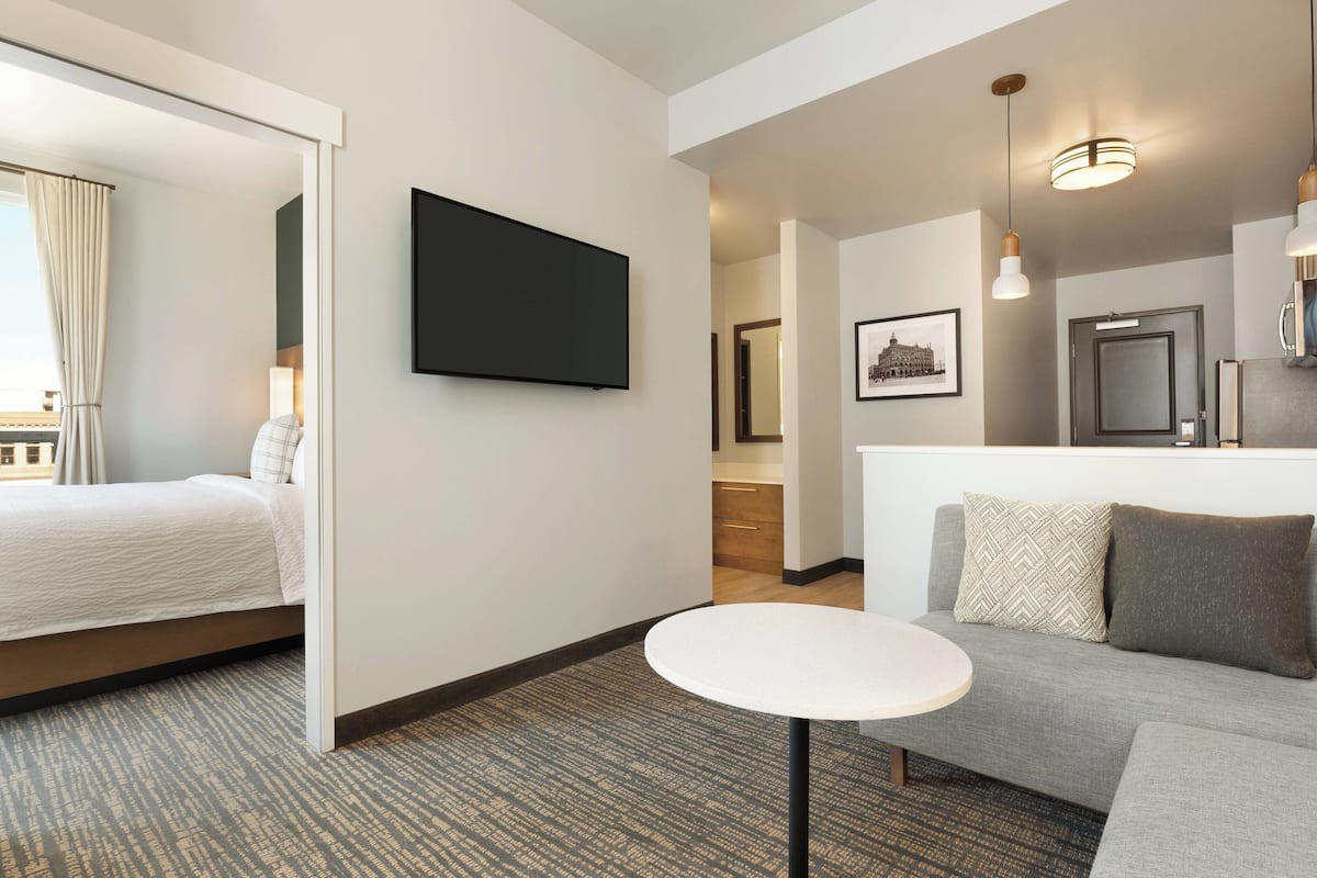 A modern hotel room with a bed, a small round table, a sofa, a TV, and a kitchenette.