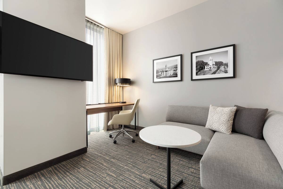 A modern hotel room with a grey sofa, a round table, a desk with a chair, and two framed pictures on the wall.