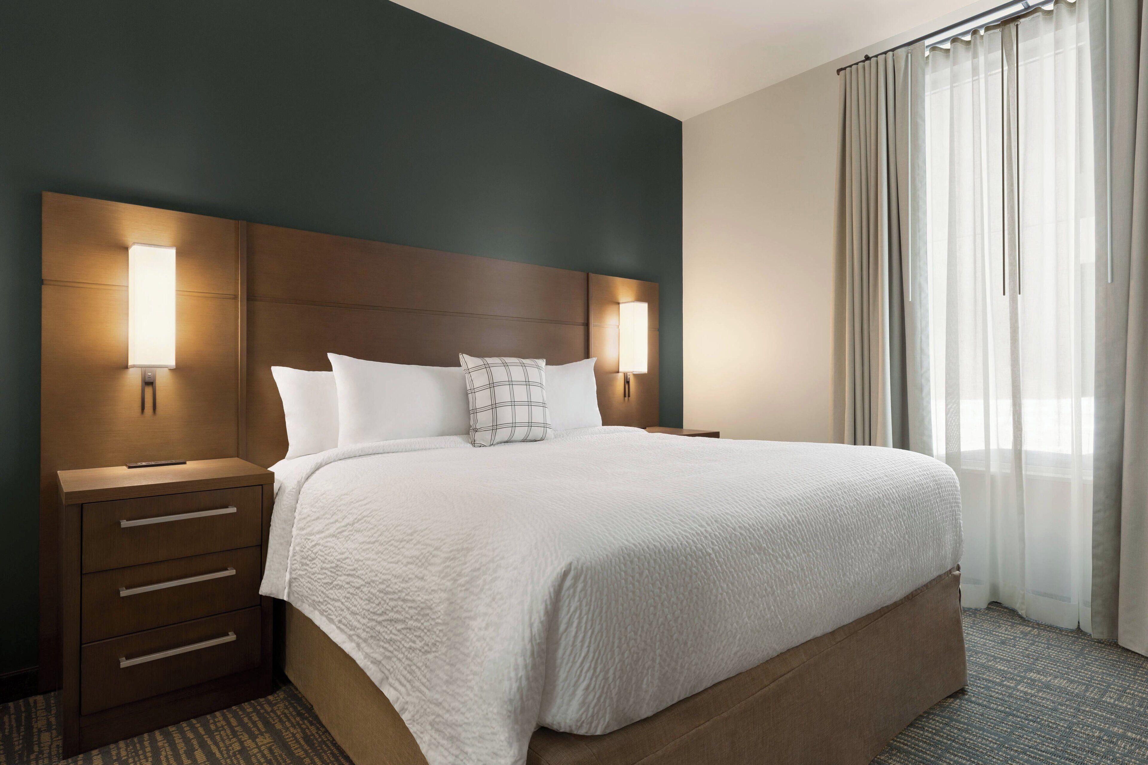 Une chambre d’hôtel avec un grand lit, deux tables de chevet avec des lampes, une tête de lit en bois et une vue sur l’extérieur à travers des rideaux transparents.