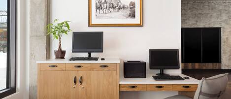 A modern office space with a wooden cabinet, two computer monitors, a desk, a chair, a framed photograph on the wall, and a potted plant.