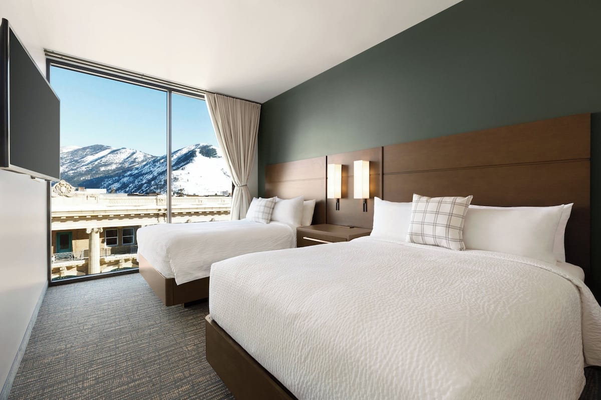 A hotel room with two beds, a large window with a mountain view, a flat-screen TV, and a wooden headboard.