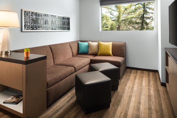 Une chambre d’hôtel moderne avec un canapé sectionnel marron, deux poufs noirs, une petite table avec une lampe, une œuvre d’art abstraite encadrée et une fenêtre donnant sur des arbres.