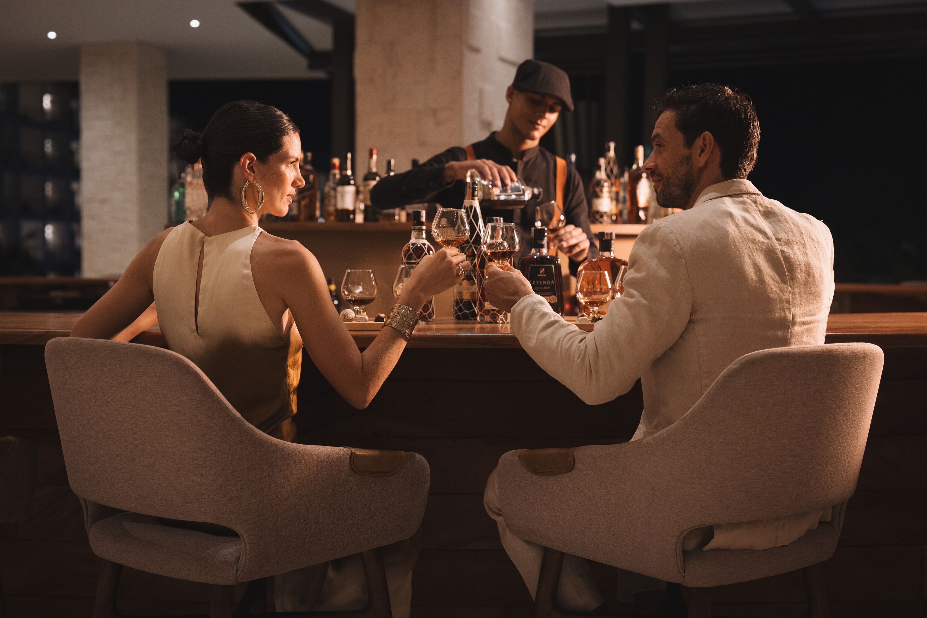 A bar setting with two people seated at a table, a bartender preparing drinks, and a variety of bottles on the bar counter.