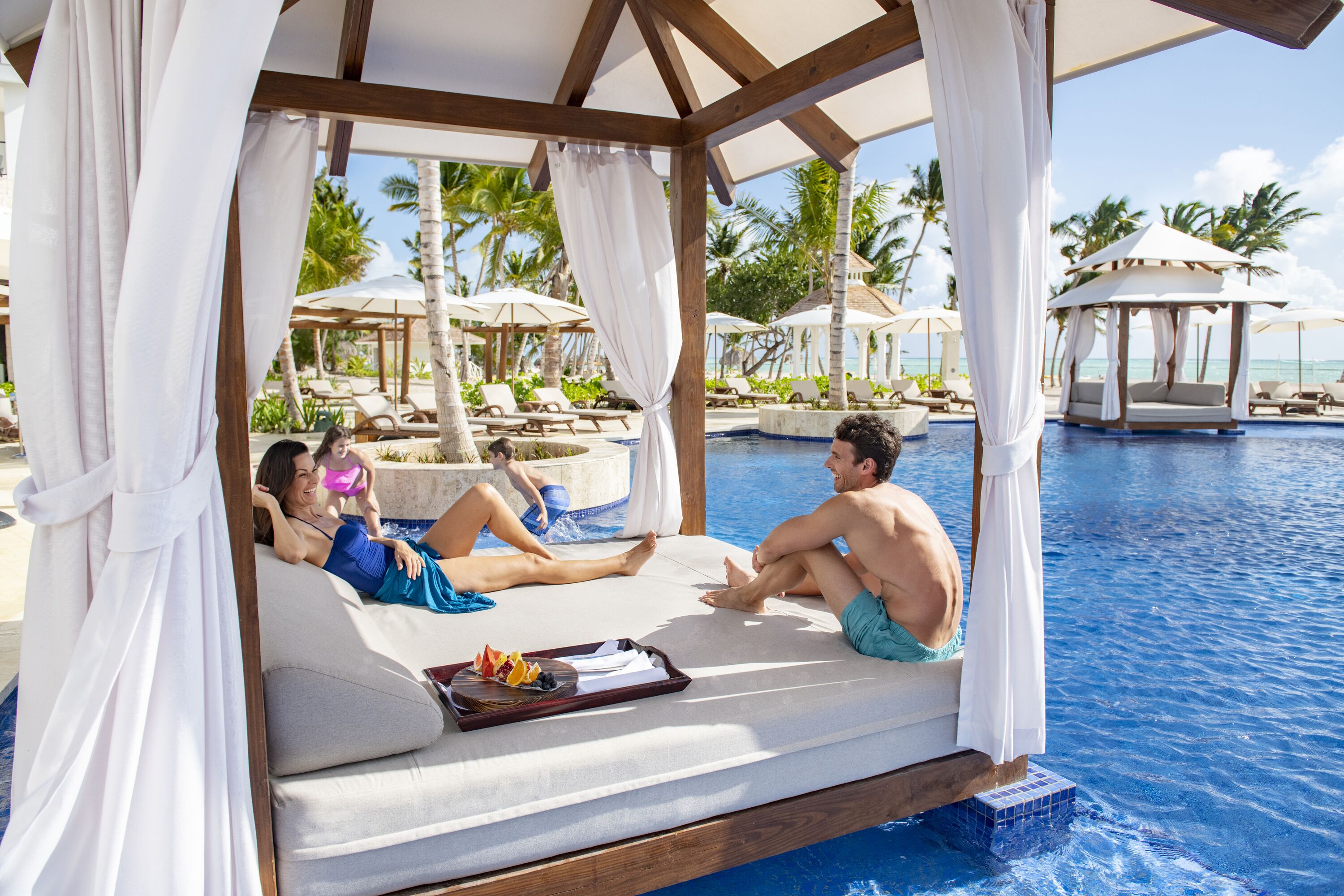 A couple relaxing on a cushioned bench by a pool, with a canopy overhead and palm trees in the background.