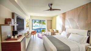 A hotel room with a bed, a desk, a TV, a view of the ocean, and a ceiling fan.