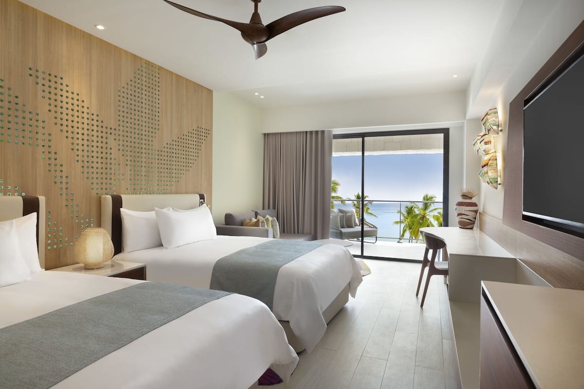 A hotel room with two beds, a desk, a chair, a TV, and a balcony with a view of the ocean.