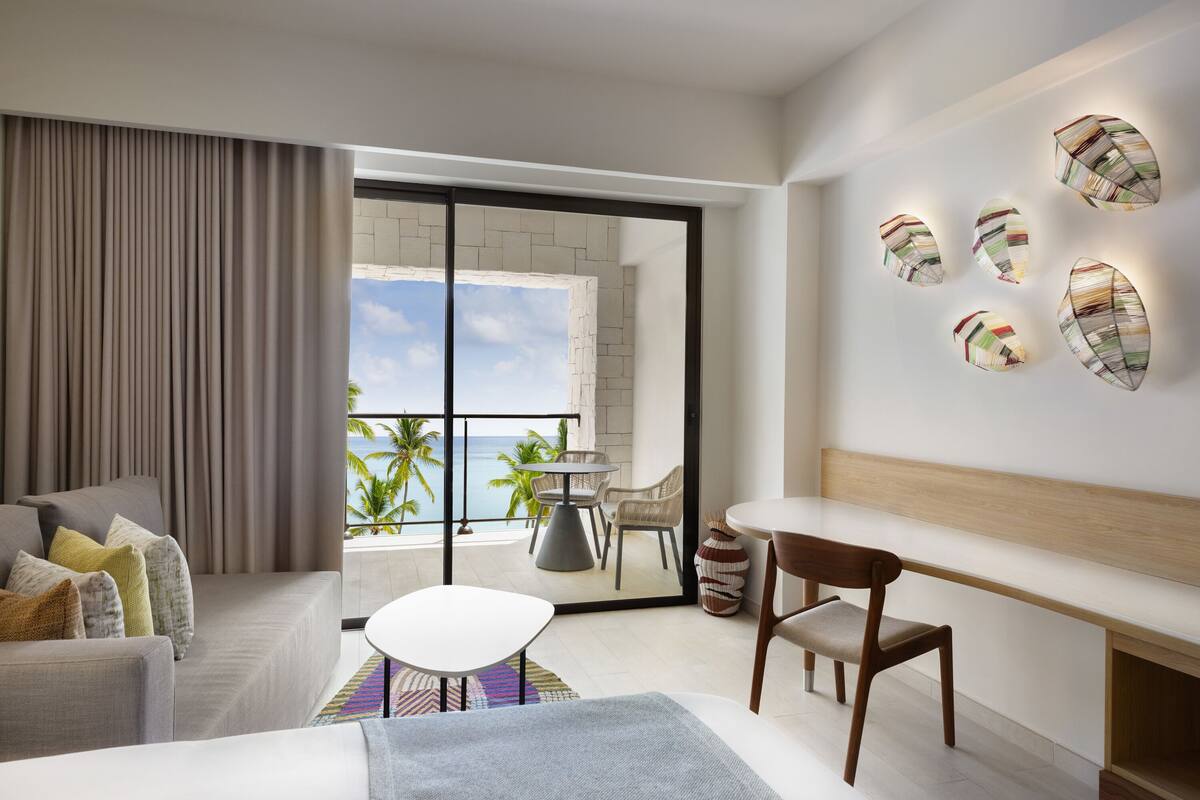 A modern hotel room with a balcony, a sofa, a dining table, and a bench.