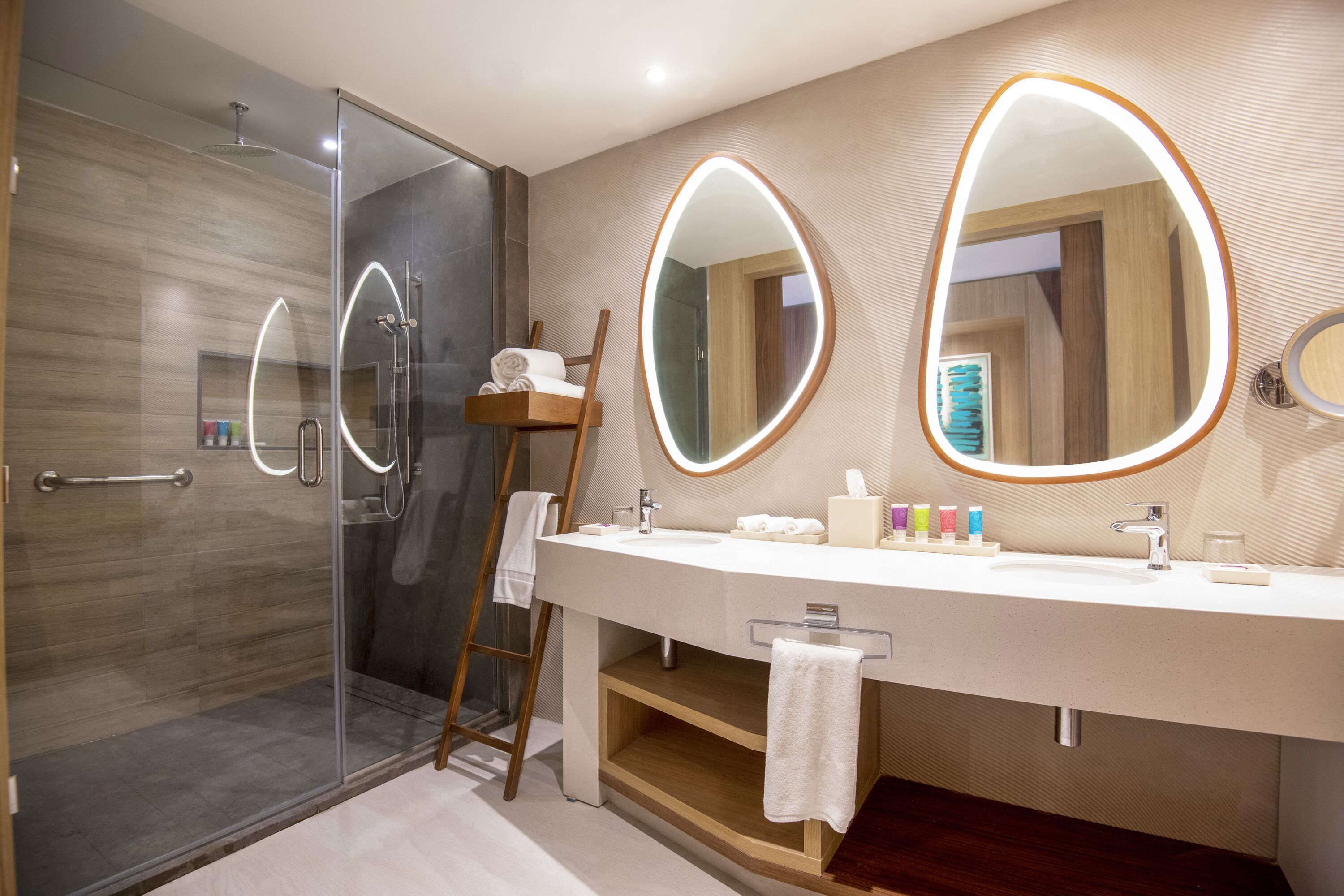 Un baño moderno con cabina de ducha de vidrio, una escalera de madera, un mueble de tocador grande con espejo y un perchero para toallas.