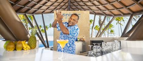 Une personne prépare une boisson dans un bar, avec des fruits tropicaux et une vue sur la plage.