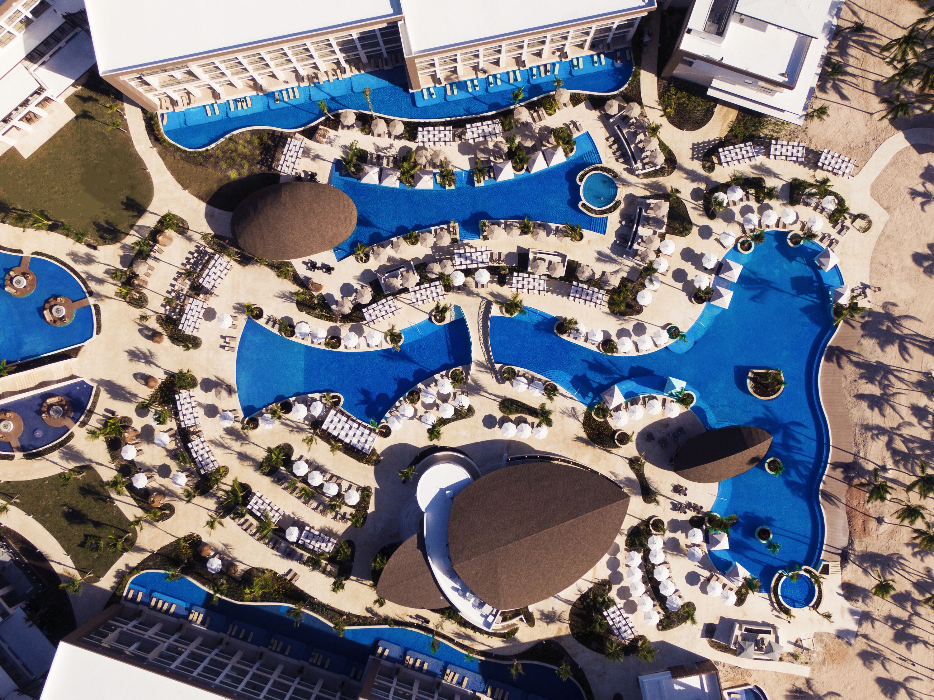 Luchtbeeld van een resort met meerdere zwembaden, ligbedden en palmbomen.