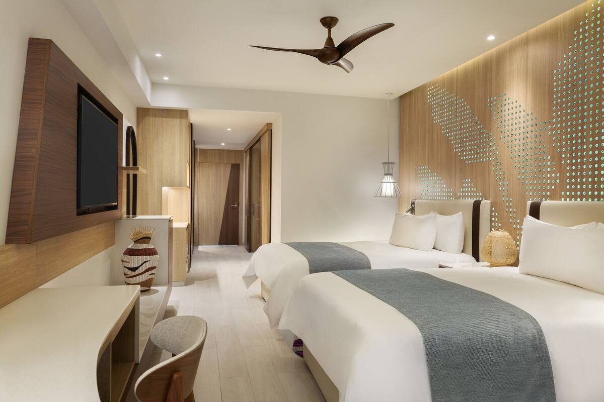 A modern hotel room with two beds, a desk, a chair, a ceiling fan, and a decorative wall.