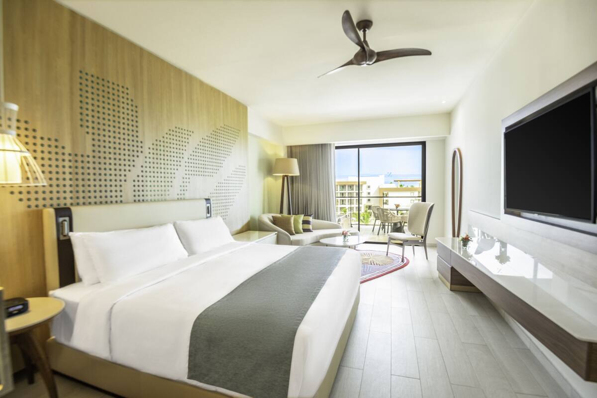 A modern hotel room with a large bed, a flat-screen TV, a ceiling fan, and a view of the city through the window.