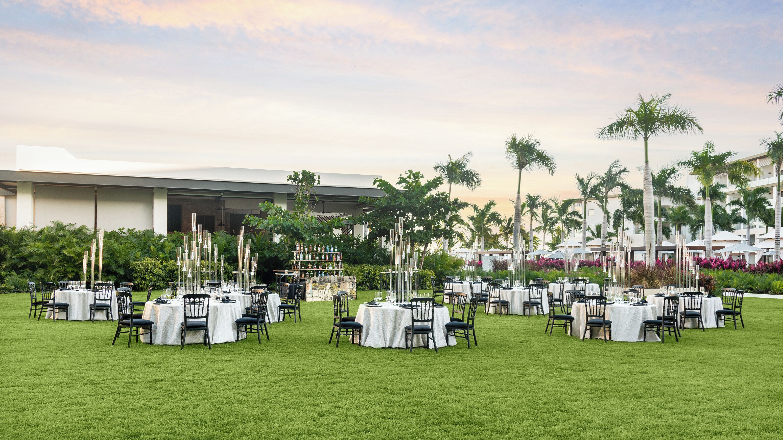 An outdoor dining area with round tables and chairs, surrounded by greenery and palm trees, with a building in the background.