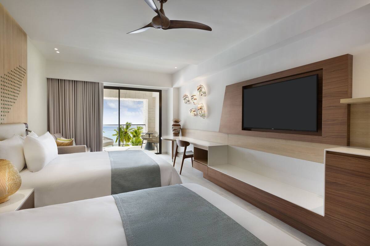 A modern hotel room with two beds, a desk, a flat-screen TV, and a large window with a view of the ocean.