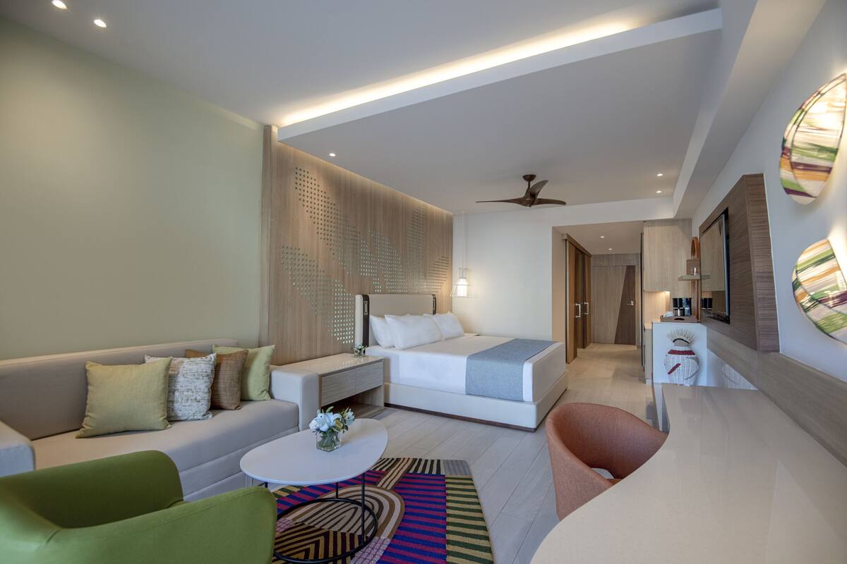 A modern hotel room with a bed, a sofa, a coffee table, and a small desk with a lamp.