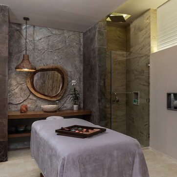 A spa room with a massage table, a wooden basin, a glass shower enclosure, and a shelf with spherical objects.