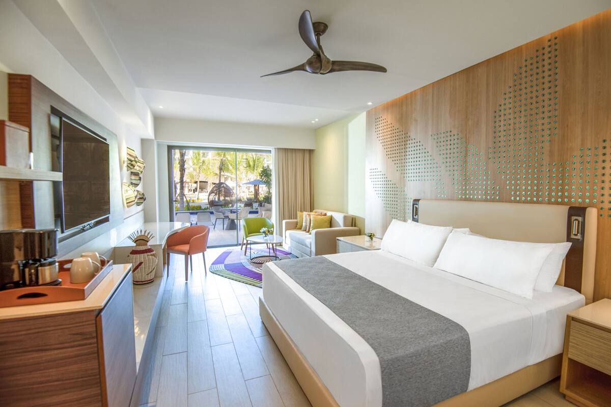 A modern hotel room with a large bed, a desk with a chair, a balcony with a view, and a ceiling fan.