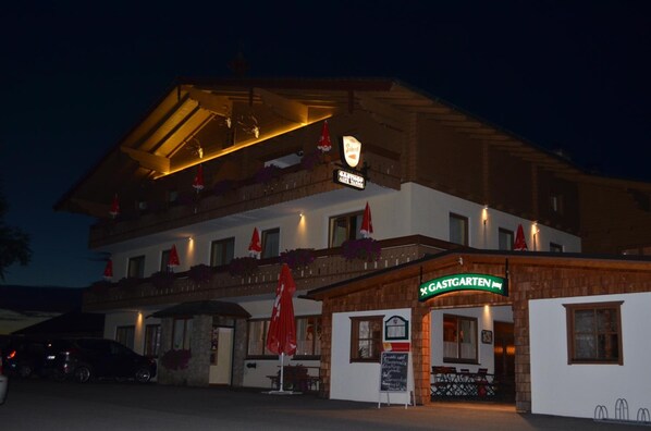 A hotel with a traditional alpine architectural style, featuring a prominent entrance with a sign, multiple balconies with red umbrellas, and a car parked in front.