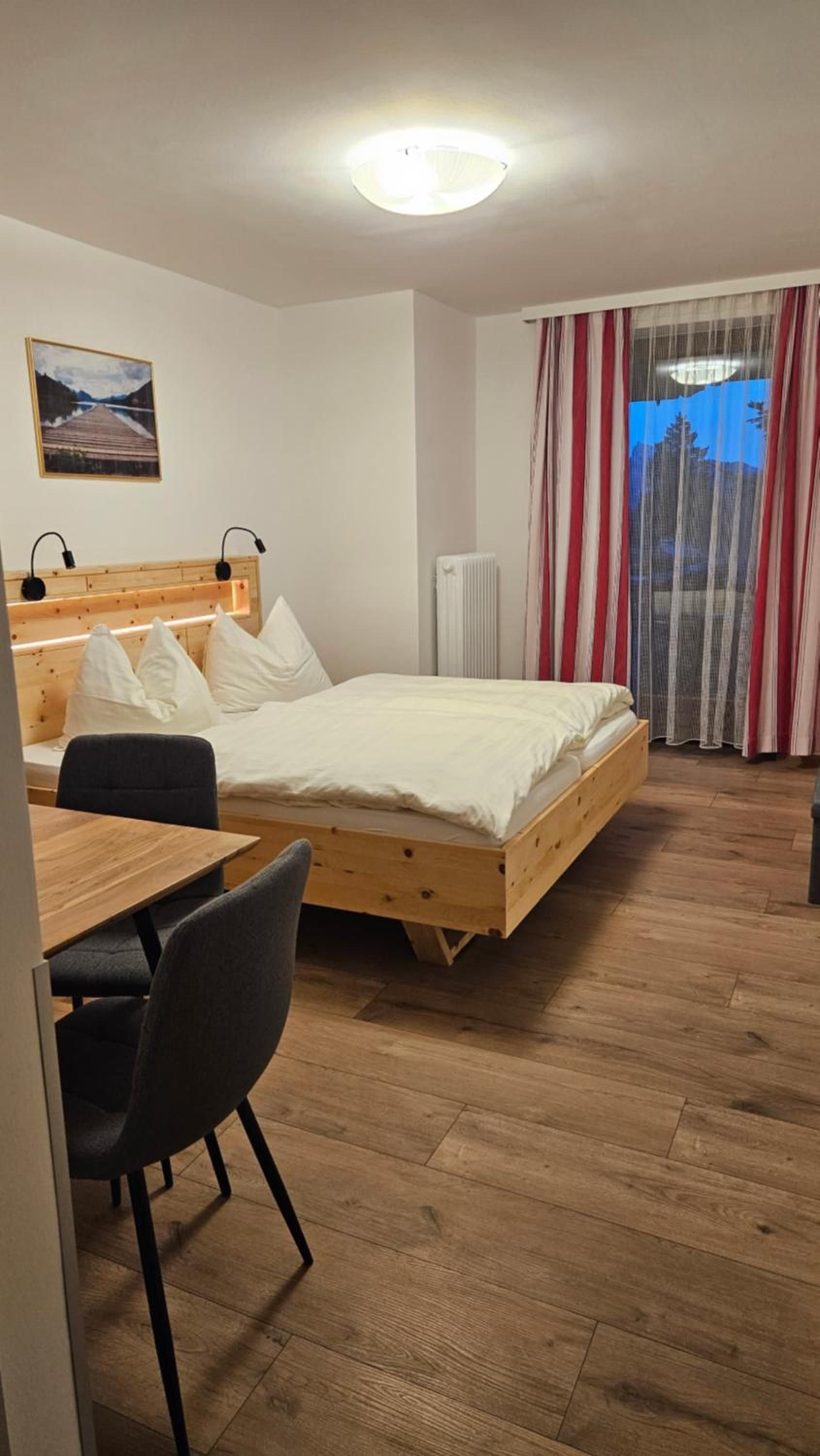 Een kamer met een bed, een bureau en een stoel. Er is een radiator en een raam met gordijnen.