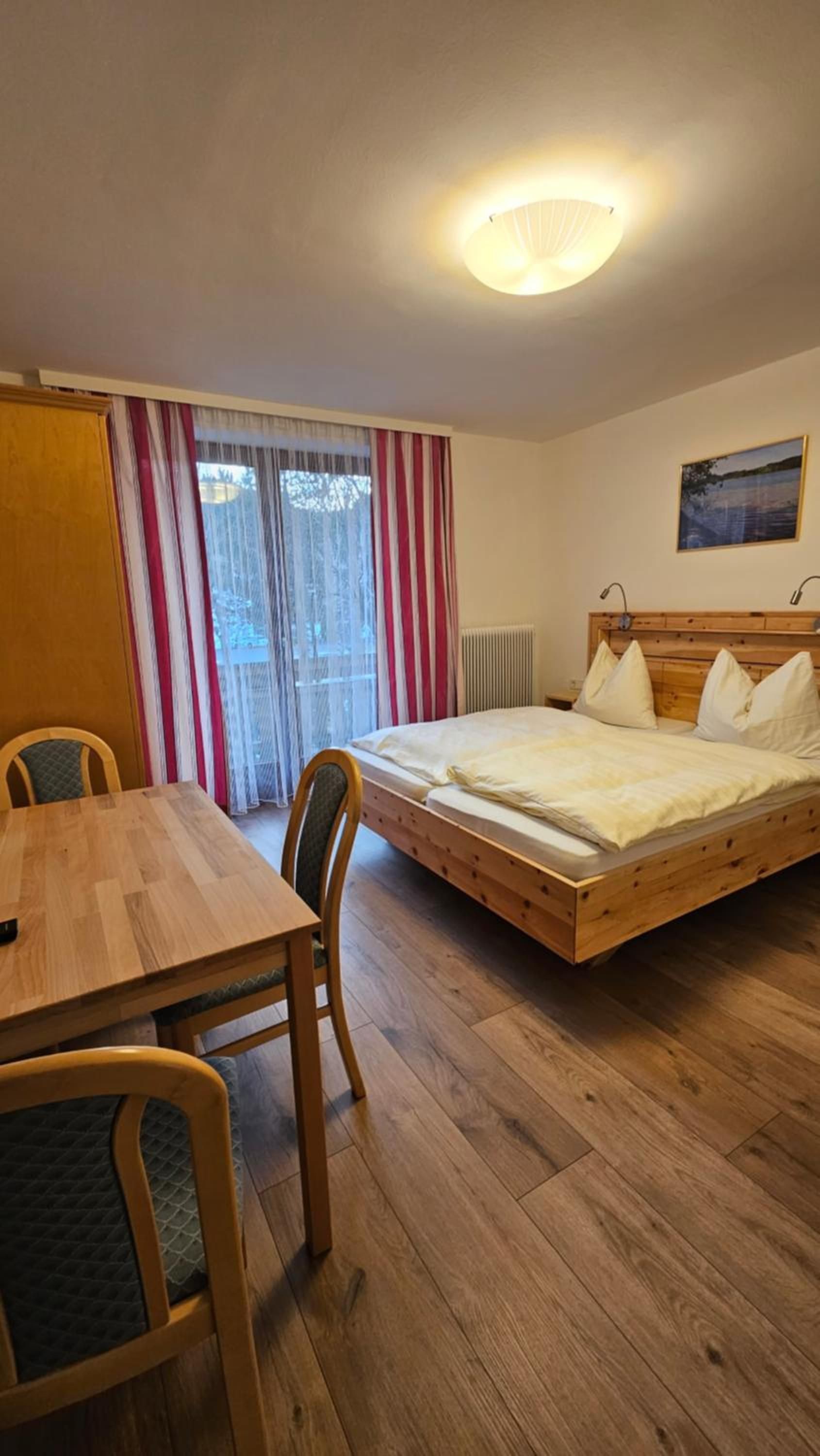 Een kamer met een houten bed, een eettafel, stoelen, een raam met gordijnen en een plafondlamp.