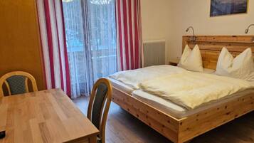 Een kamer met een houten bed, een eettafel, stoelen, een raam met gordijnen en een plafondlamp.