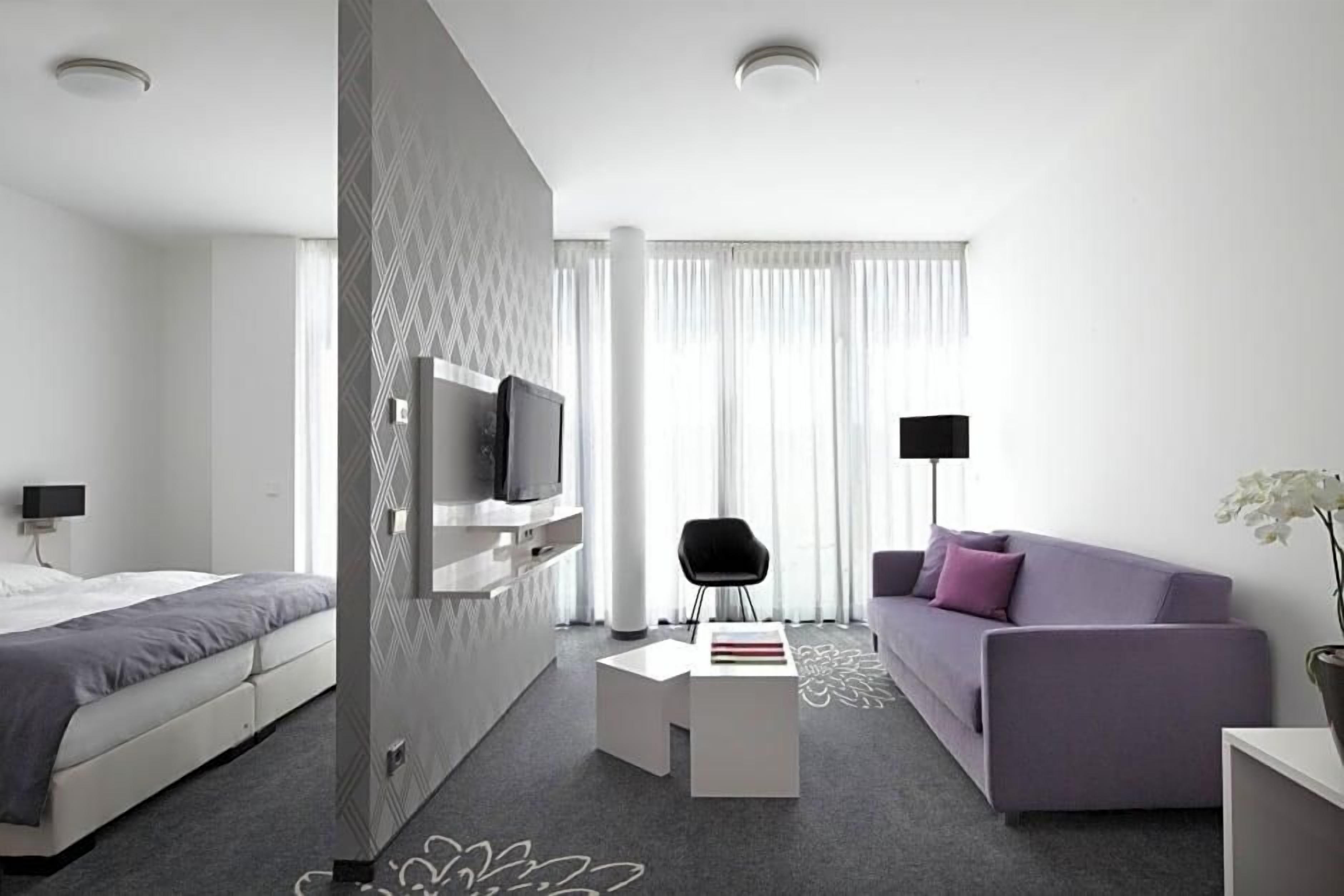 Ein modernes Hotelzimmer mit Bett, einer Couch, einem Fernseher, einem kleinen Tisch und einem Sessel.