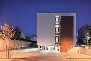 Een modern gebouw met een platte gevel, veel ramen en een parkeerplaats met enkele auto's.