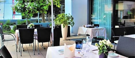Un espacio de comedor al aire libre con mesas dispuestas para dos, sillas y un paraguas rojo.