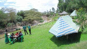 A group of people sitting at a table outdoors, surrounded by greenery and a river. There is a striped canopy structure nearby.