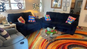 A living room with two dark sofas, colorful cushions, a glass coffee table with a fish decoration, and a vibrant striped rug.