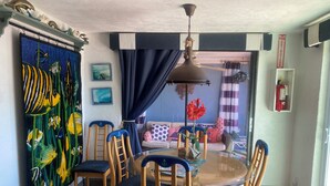 A dining area with a round glass table, wooden chairs with blue cushions, a ceiling fan, and a fish-themed wall hanging.