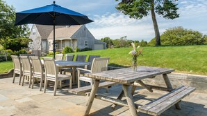 An outdoor patio with a wooden table and chairs, a blue umbrella, and a house in the background. - Nantucket Private Oasis - 6BR Pet-Friendly Home, Short Drive to Quidnet Beach (Nantucket)