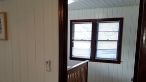 Ein Zimmer mit Holzboden, einem Holzstuhl, einem Fenster mit Jalousien und einer Tür zu einem weiteren Raum.