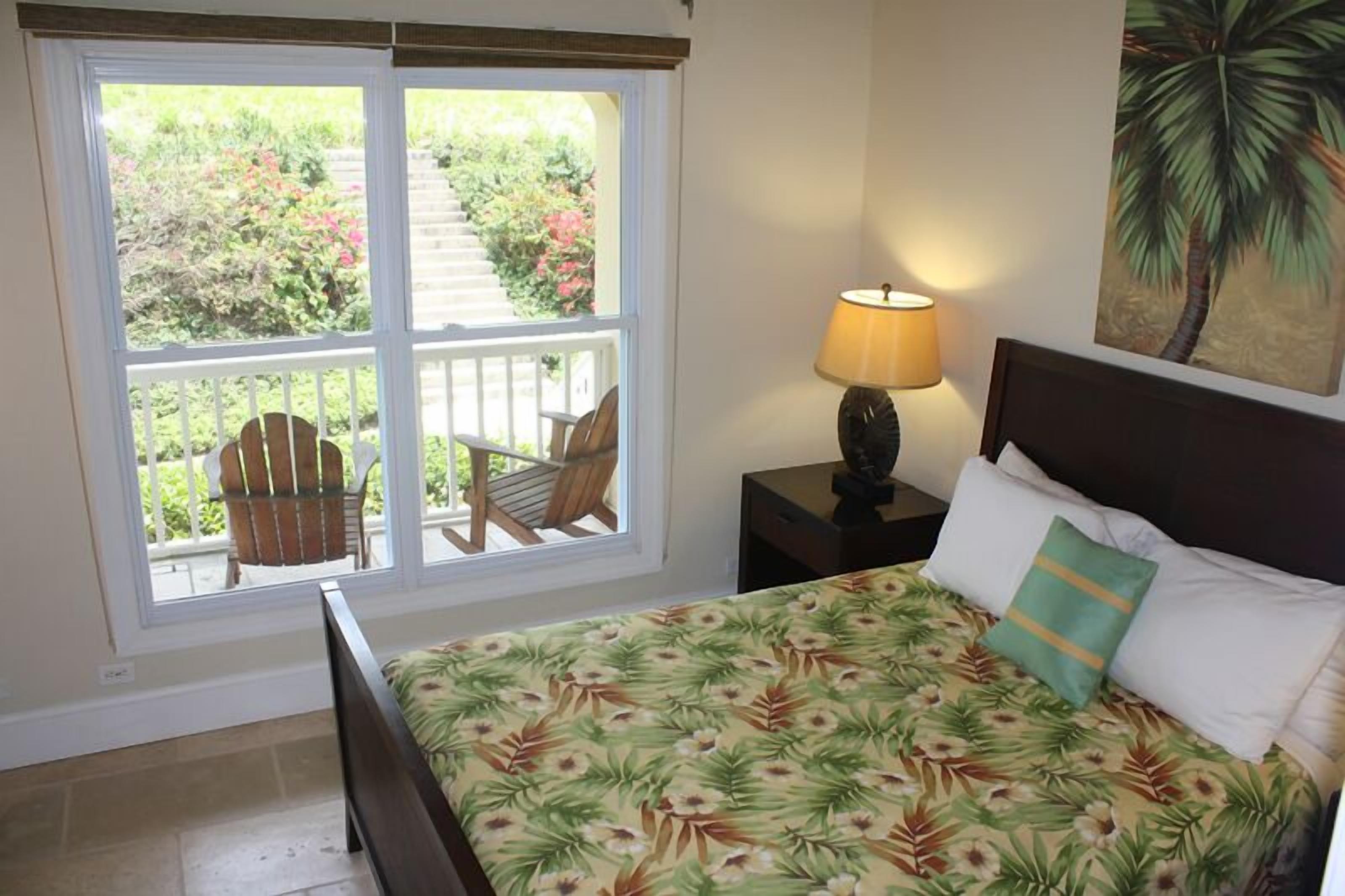 Une chambre à coucher avec un lit, une table de chevet avec une lampe, une fenêtre donnant sur un balcon et un jardin.