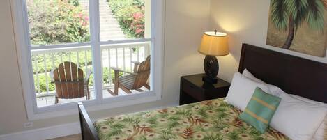 Une chambre à coucher avec un lit, une table de chevet avec une lampe, une fenêtre donnant sur un balcon et un jardin.