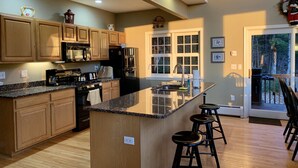 A kitchen with wooden cabinets, a granite countertop, a central island with stools, a refrigerator, a stove, and a window with a view of trees.