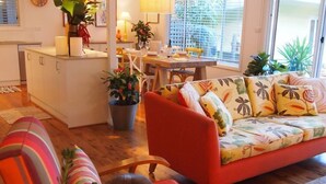 A cozy living room with a sofa, a chair, and a cat. There is a dining table with chairs, a kitchen area, and a large window with plants. - Retro Beach House on Pearl Beach (Pearl Beach)