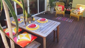 An outdoor patio with a wooden table set for dining, surrounded by wicker chairs and a potted plant. - Retro Beach House on Pearl Beach (Pearl Beach)