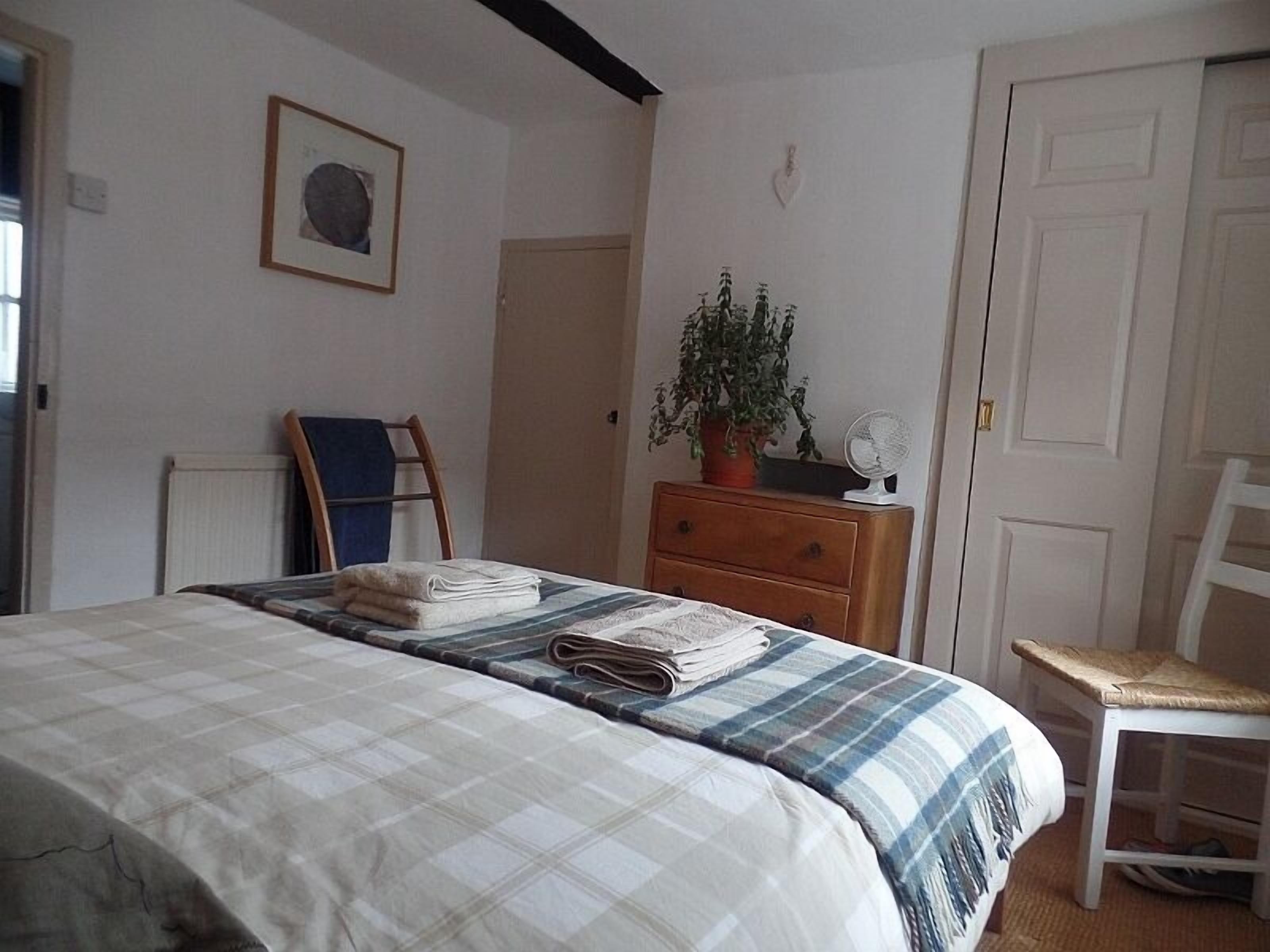Un dormitorio con cama, cómoda, silla, planta en maceta, ventilador y puerta.