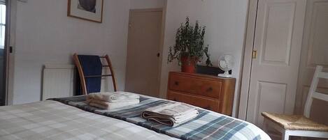 Un dormitorio con cama, cómoda, silla, planta en maceta, ventilador y puerta.